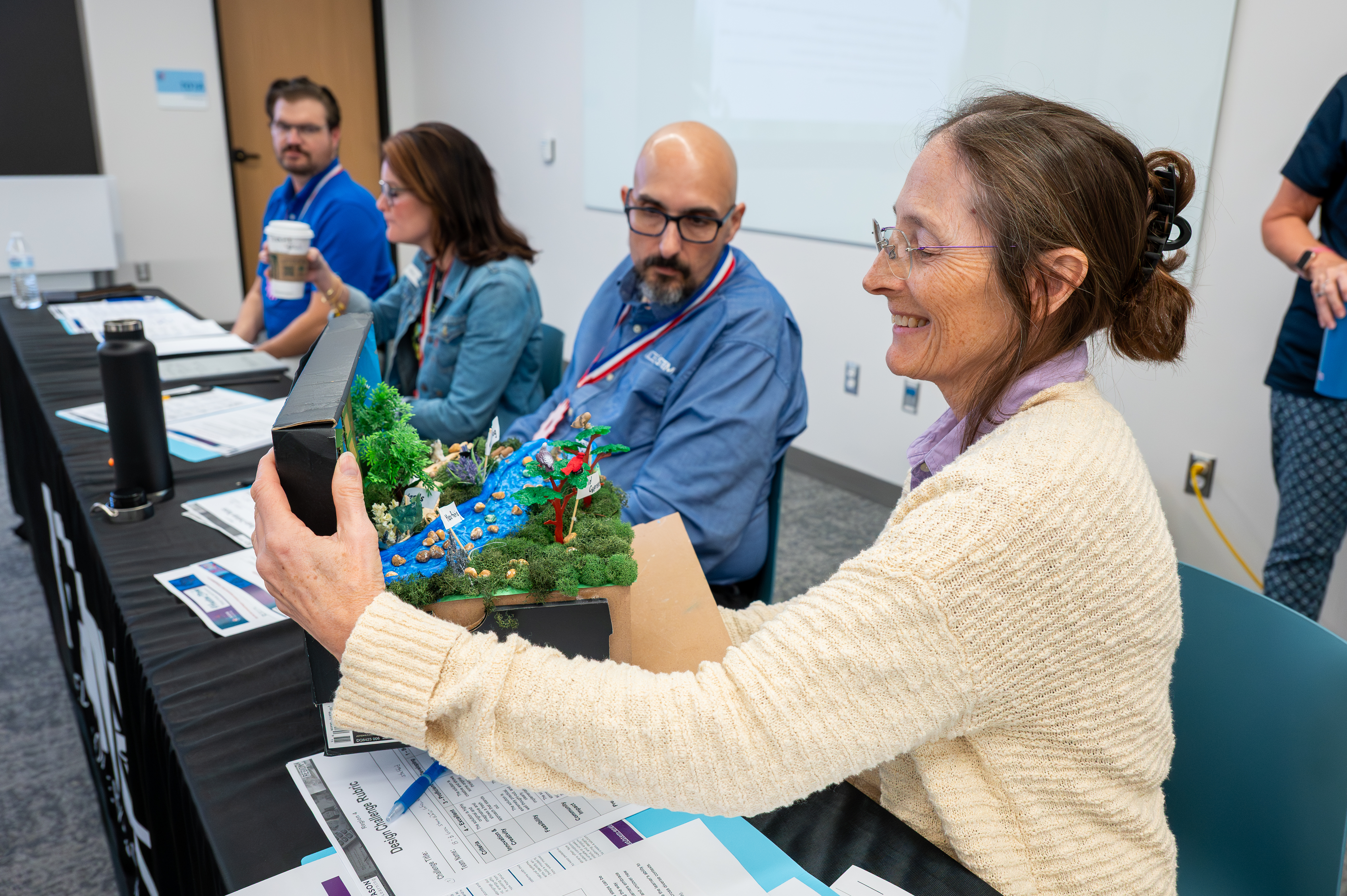 A woman in a pink shirt demonstrating a small engineering model or activity to two colleagues seated across a table during a workshop.