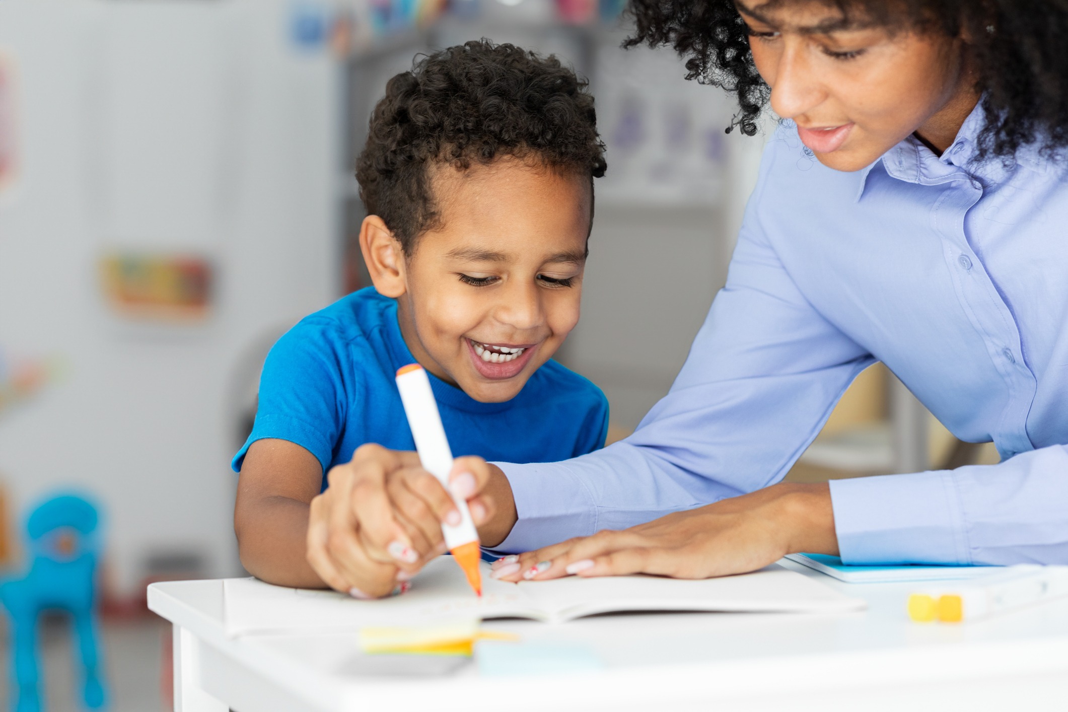 School teacher working with positive black schoolboy, sitting at table and learn writing in classroom interior