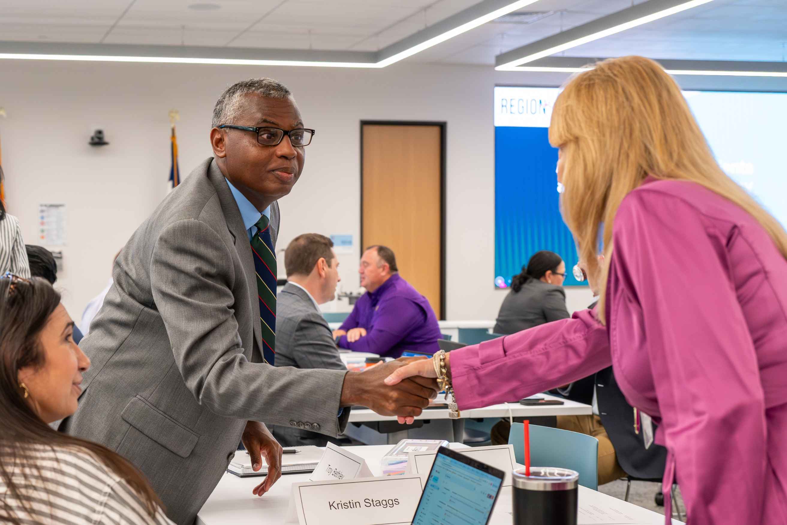 Two professionals in business attire shaking hands across a meeting table during a workshop session.